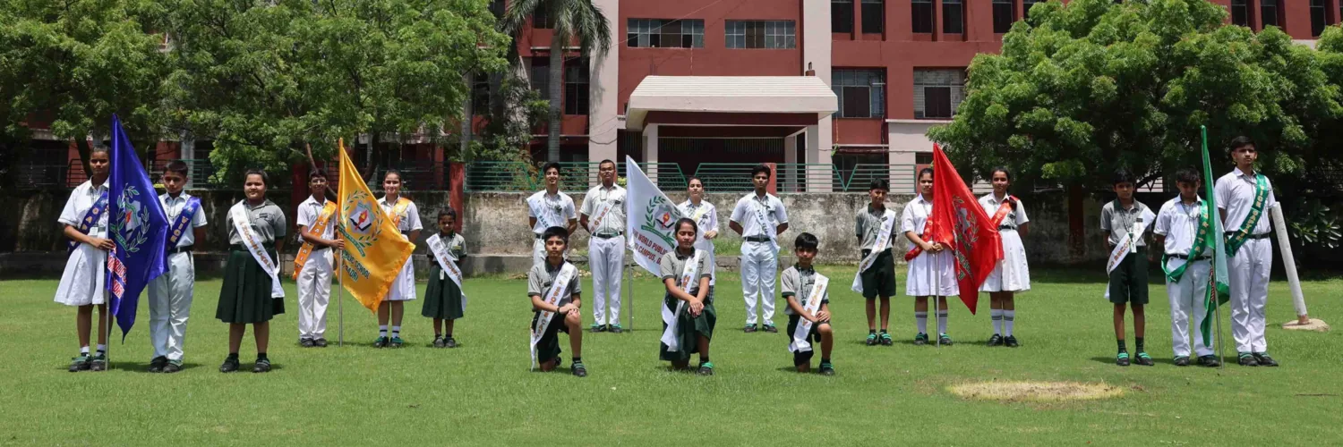 Students holding house flags on the school ground, with the Delhi World Public School building behind them